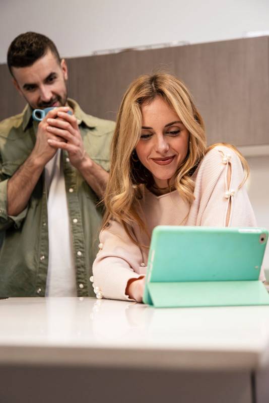 Smiling happy man drinking coffee and woman using a touch pad in a video call. Lovely handsome couple looking tablet together in the kitchen.