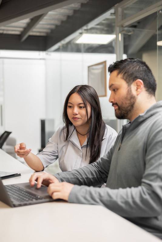 woman and a man stand side by side, looking at a laptop screen and discussing data. The office environment