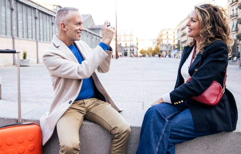 Senior tourist couple taking picture on vintage camera sitting in a bench. Mid adult man taking a photography of her wife during romantic trip outdoor.