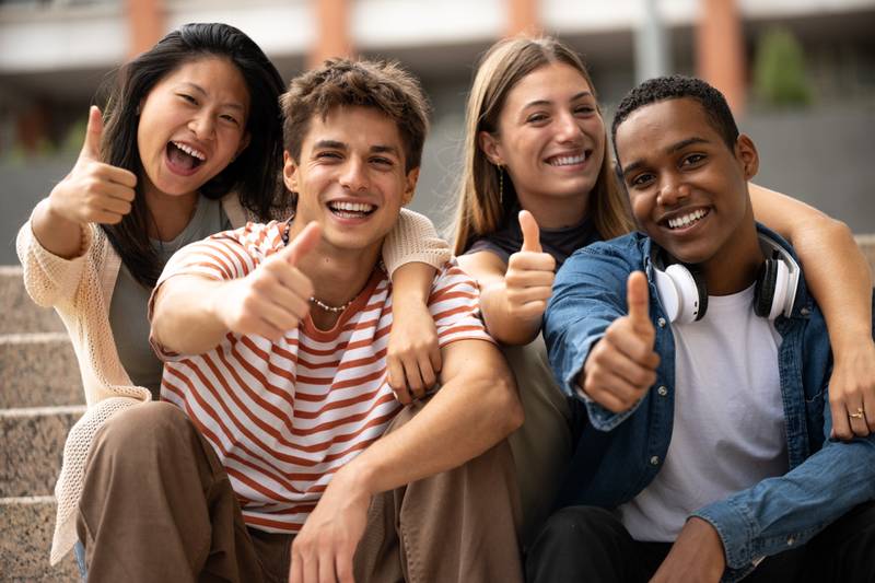Cheerful multicultural group of friends showing thumbs up sitting on stairs. Happy young students smiling and satisfied sitting outside.