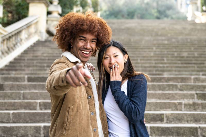 Lovely multiracial couple surprised and pointing to the camera in a park.Two happy diverse young persons smiling and pointing to the front with finger.