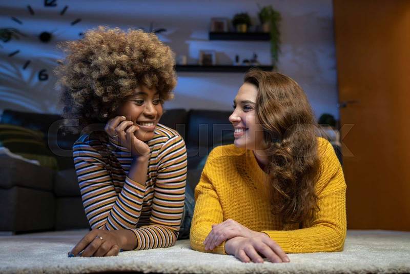 Happy multiracial lesbian couple looking each other. Portrait of a beautiful young couple lying on the floor of a living room. Bonding concept