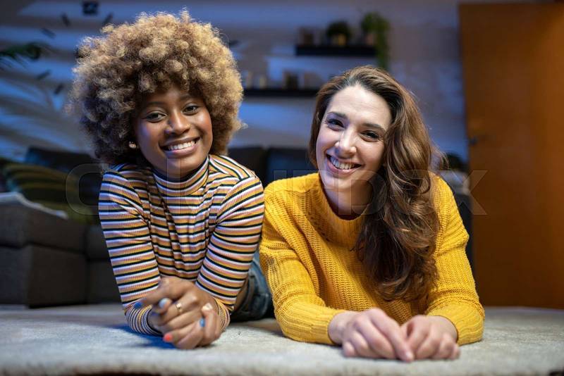 Multiracial smiling lesbian couple looking at camera. Portrait of two cheerful woman lying on the floor of a living room.