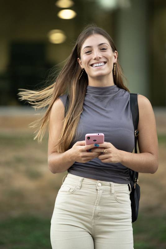 Beautiful satisfied young lady looking at camera texting and standing outside.Joyful confident blond woman staring at camera in the street while using her phone.