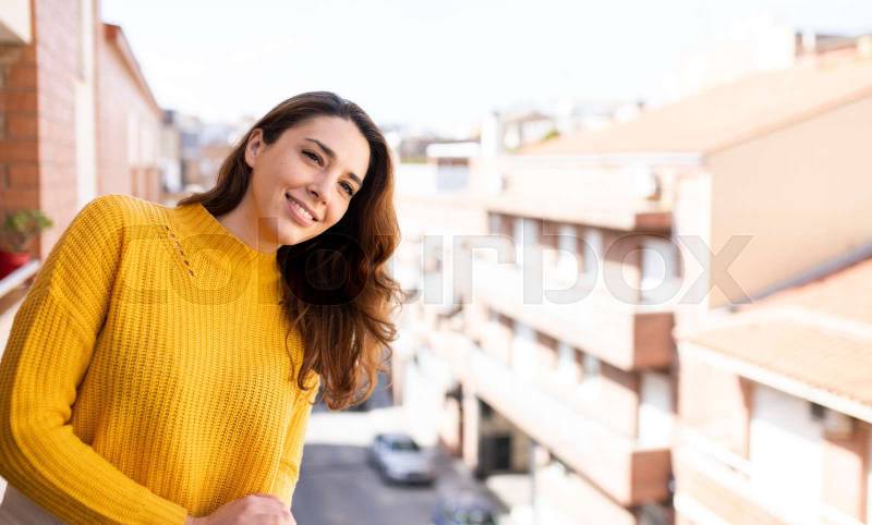 Portrait of young smiling woman looking away standing with a city background. Cheerful female looking satisfied on a balcony.