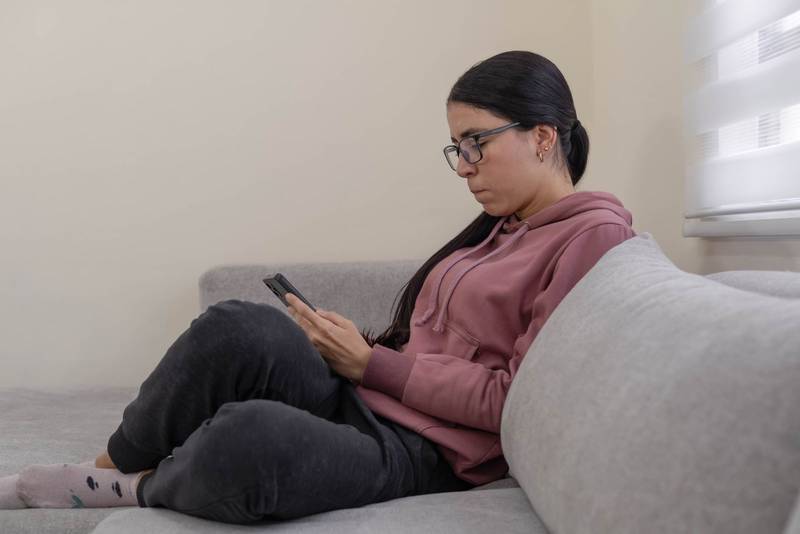 woman in glasses and pink hoodie sitting comfortably on sofa, talking and smiling during a pleasant mobile phone conversation