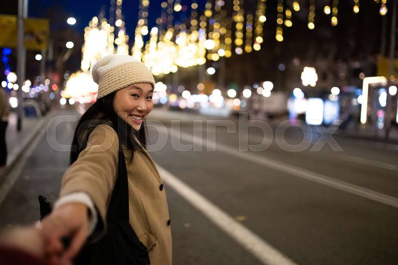 Beautiful woman holding hand in the street with christmas lights. Cheerful lady with warm hat looking at camera in winter.