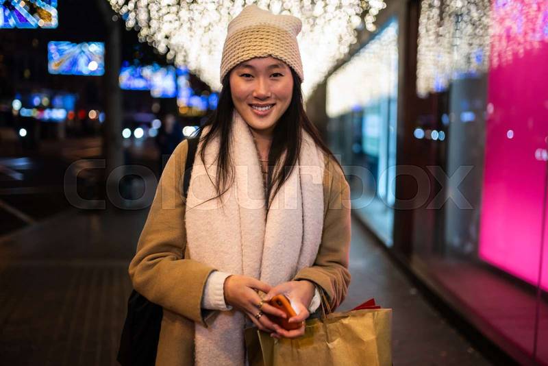 Young smiling girl looking at camera in the street at night. Happy woman wearing warm hat standing outstide in winter.