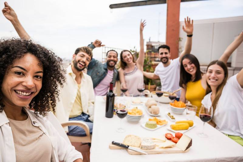 Cheerful diverse group of friends having fun and dinner in a rooftop. Multiracial young people taking a picture in a outdoor table while eating and drinking. Focus on girl.