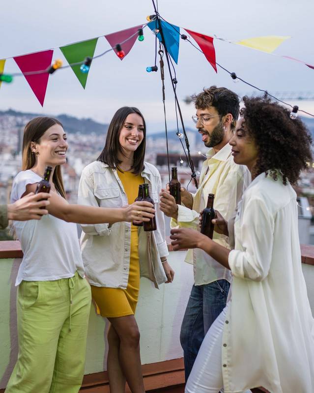 Diverse group of friends having fun and drinking beer in a rooftop party. Multiracial people toasting with bottles at evening in a terrace