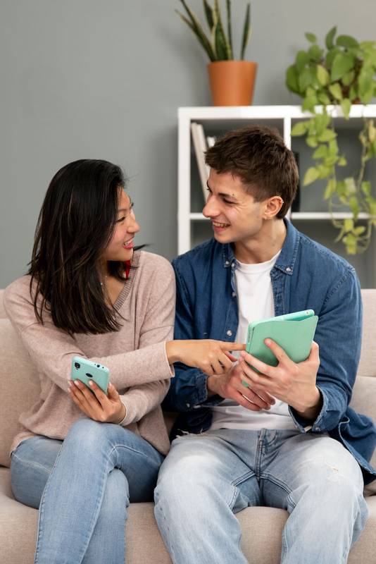 Diverse young couple smiling and having fun using online device. Smiling woman pointing tablet and man sitting on a couch in their living room at home.