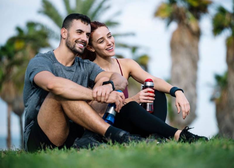 Exhausted young adult couple resting in the floor after running together in the park. Affectionate satisfied athletic male and female sitting in the grass in sportswear training and workout outside. 
