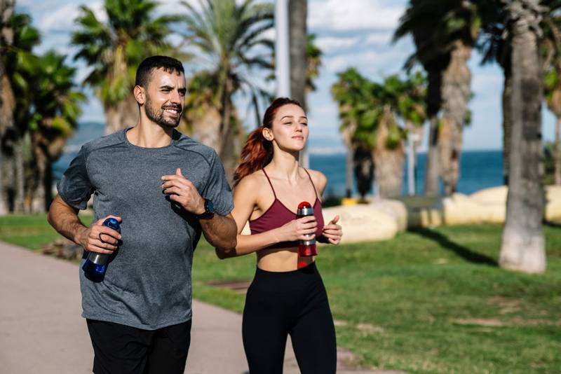 Young adult couple running together holding water bottles while jogging. Happy athletic male and female in sportswear training with drinking workout outside.