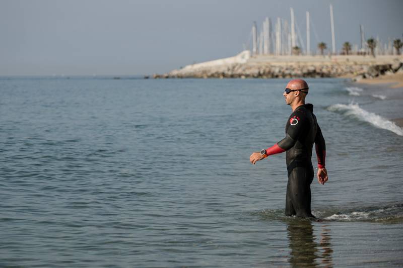 Swimmer wearing neoprene entering the water in an urban beach. Athlete ready for swimming in the ocean with wetsuit and glasses.