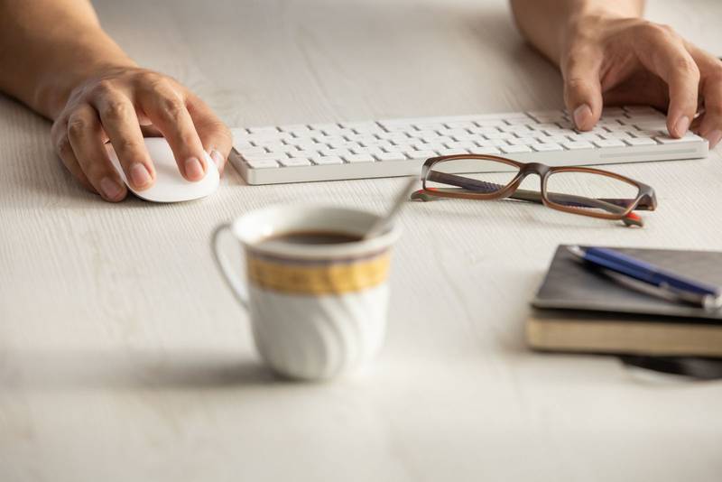 person working at his desk, detail of his hands next to a modern keyboard, glasses, pen, cup
