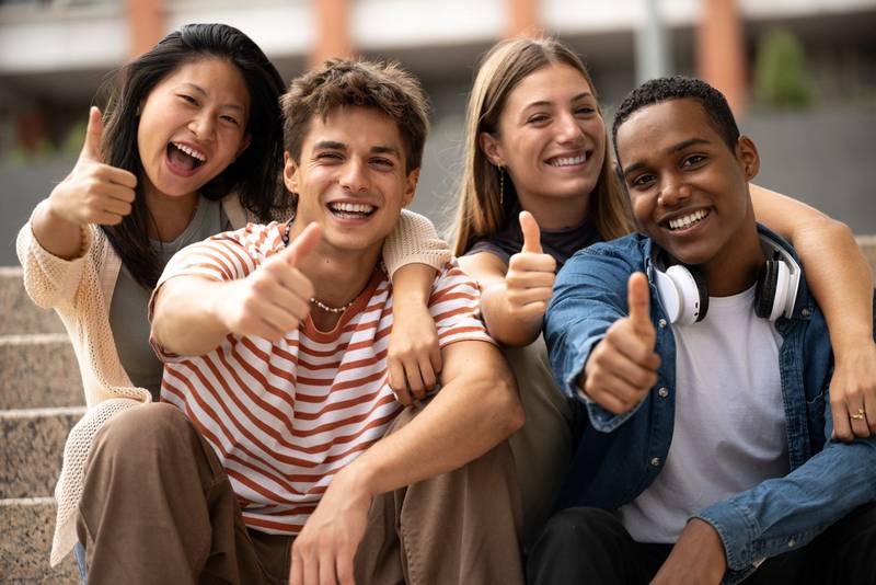 Cheerful multicultural group of friends showing thumbs up sitting on stairs. Happy young students smiling and satisfied sitting outside.