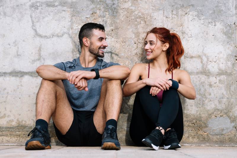 Exhausted young adult couple resting in the floor after running together in the park. Affectionate satisfied athletic male and female sitting in the grass in sportswear training and workout outside. 