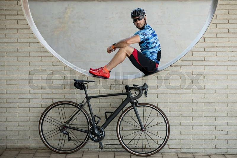 Fit man in sport clothes sitting in a wall with a bike looking at camera. Athletic cyclist resting next to his cycle.