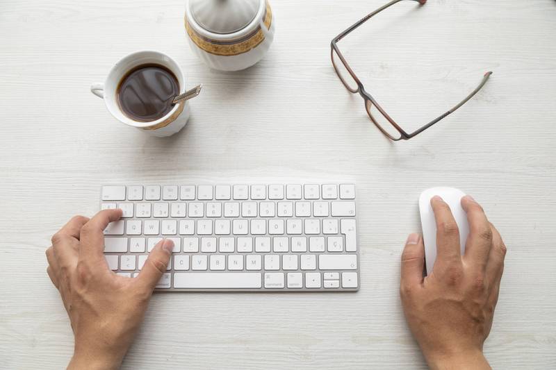 hands of a person typing on a modern keyboard and using a mouse, around them a pair of glasses, a cup of coffee