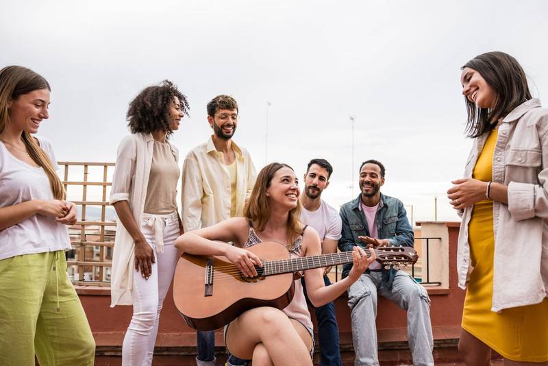 Beautiful young lady playing guitar with friends in a rooftop party. Happy carefree girl playing music in a terrace with cheerful people around.