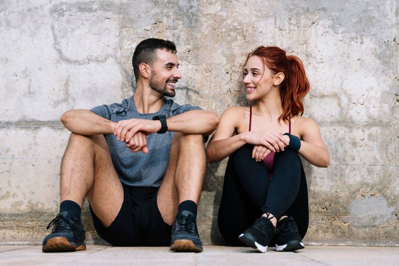 Exhausted young adult couple resting in the floor after running together in the park. Affectionate satisfied athletic male and female sitting in the grass in sportswear training and workout outside. 