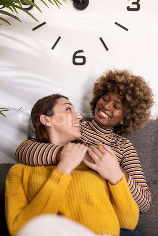 Multiracial lesbian couple hugging and laughing and relaxing in a living room. Smiling gay females couple having fun at home embracing each other on a sofa.