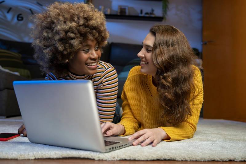 Happy female couple looking each other with a computer lying on the floor. Two lesbian woman smiling while using a laptop on carpet.