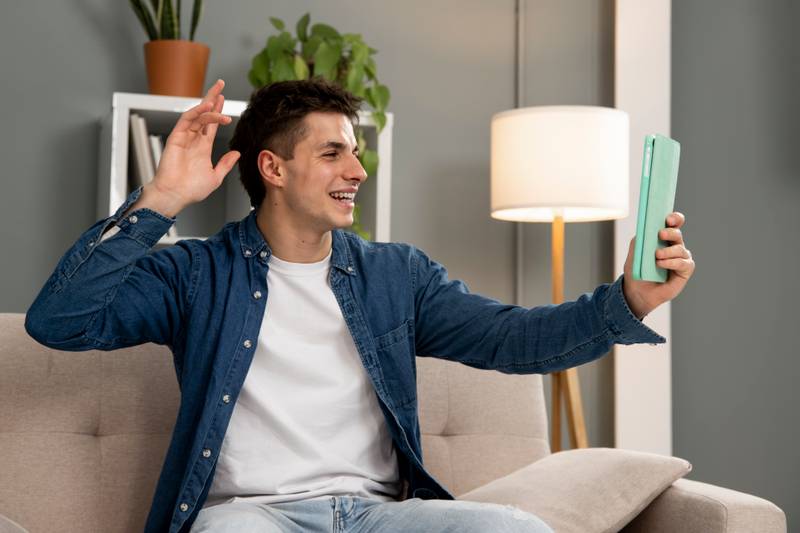 Handsome relaxed male waving in a video call with touchpad while sitting on a couch. Happy smiling young carefree man using tablet at home. 