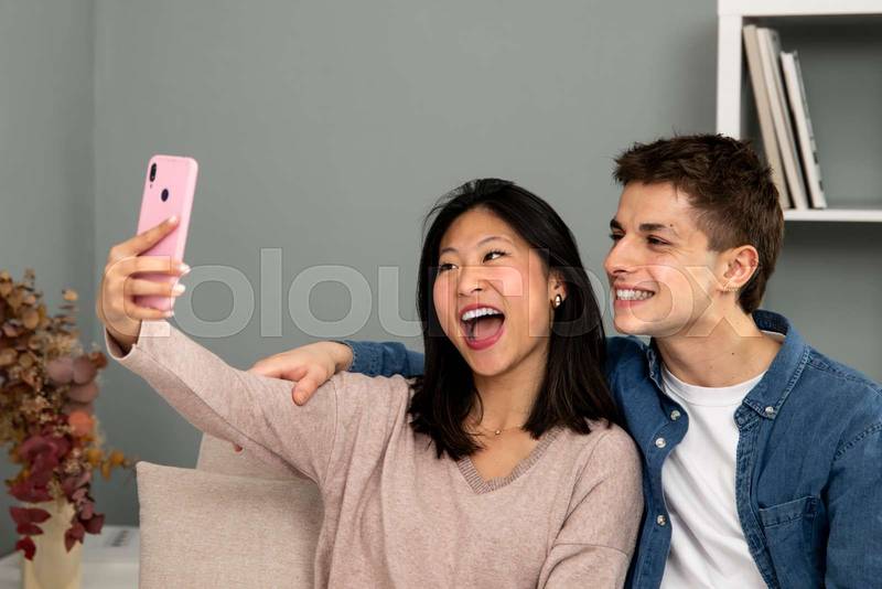 Cheerful man and woman laughing in a video call with smart phone. Diverse young couple on the sofa in their living room taking a selfie waving. 