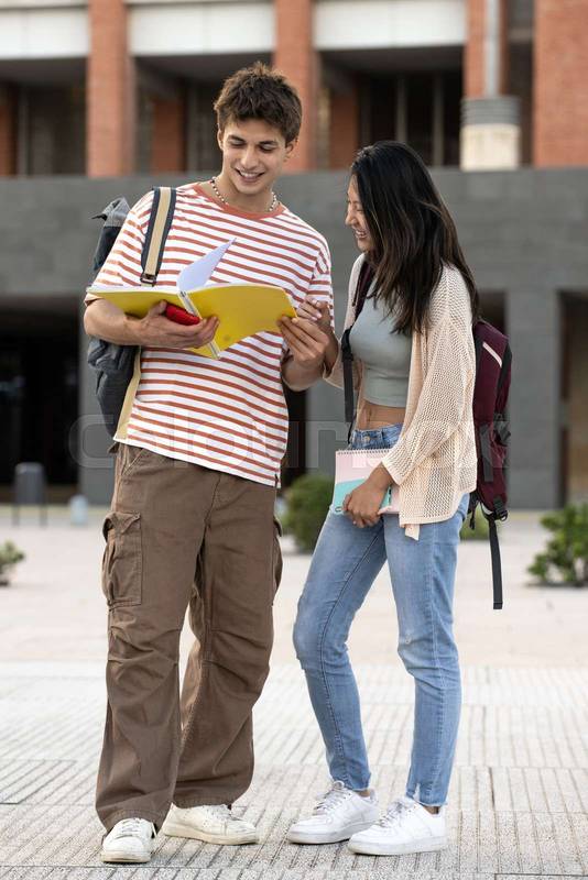 Diverse couple standing and reading notes in a campus. Two young students looking notebook smiling and studying in the street.