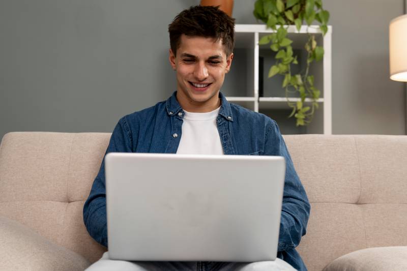 Handsome relaxed male student working with laptop while sitting on the couch. Smiling young carefree man using computer at home.