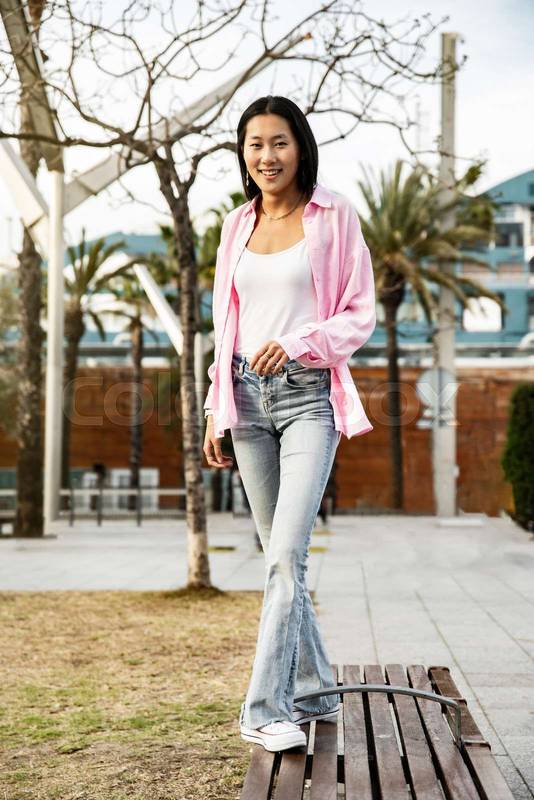 Young smiling woman balancing on bench in the street looking at camera. Carefree and confident lady walking on bench park staring at camera in a city.