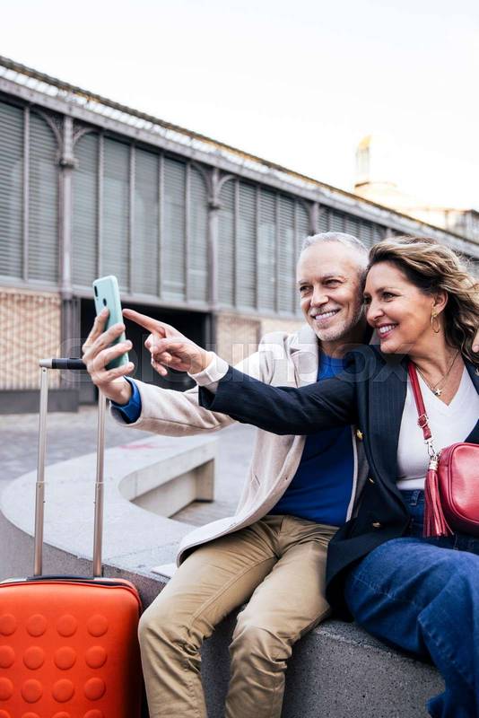 Cheerful mid adult tourist couple travelling together taking selfie sitting outdoors with luggage. Affectionate senior husband and wife taking picture in a street bench during weekend romantic trip.