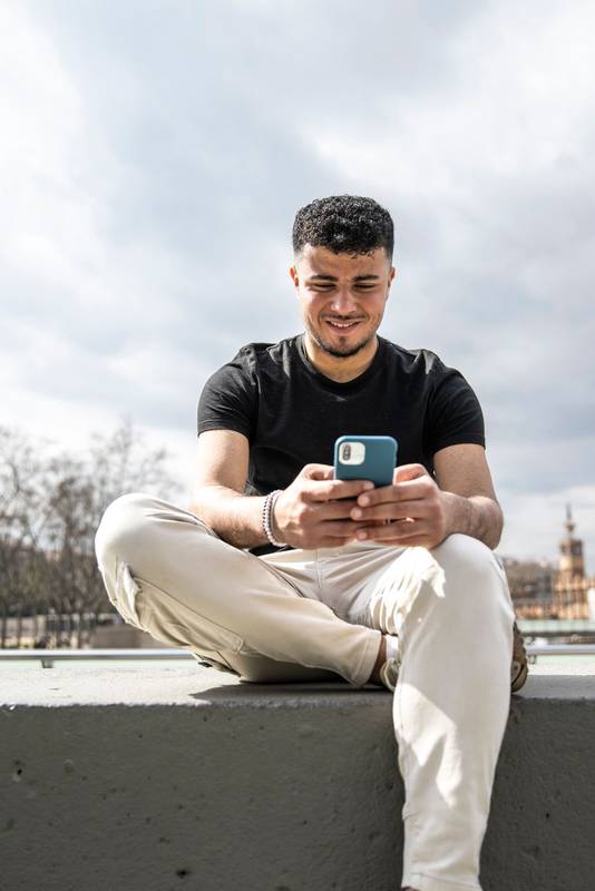 Young handsome man sitting on bank relaxed and texting with her phone. Satisfied guy smiling and using an smartphone sitting in the street.