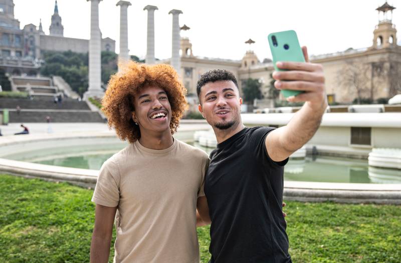 Two happy men taking a selfie with phone in park. Cheerful masculine couple smiling and taking a picture outside.