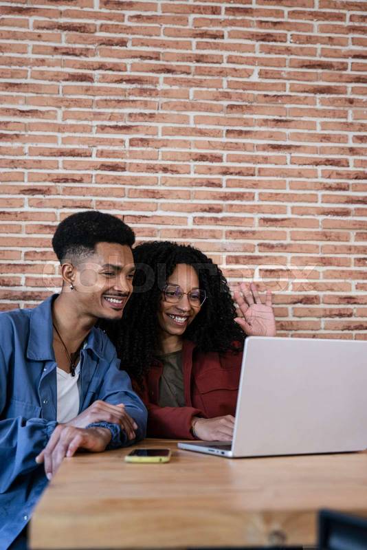 Happy young diverse couple sitting at living room having laptop video call at home. Friendly young adult woman and man in love waving at computer screen.