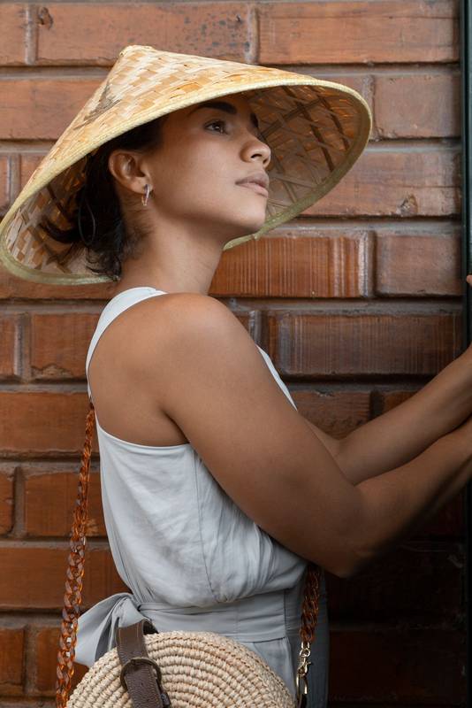 young brunette woman smiling and pointing to a window next to a brick wall, wearing a straw hat and white summer blouse, fashion