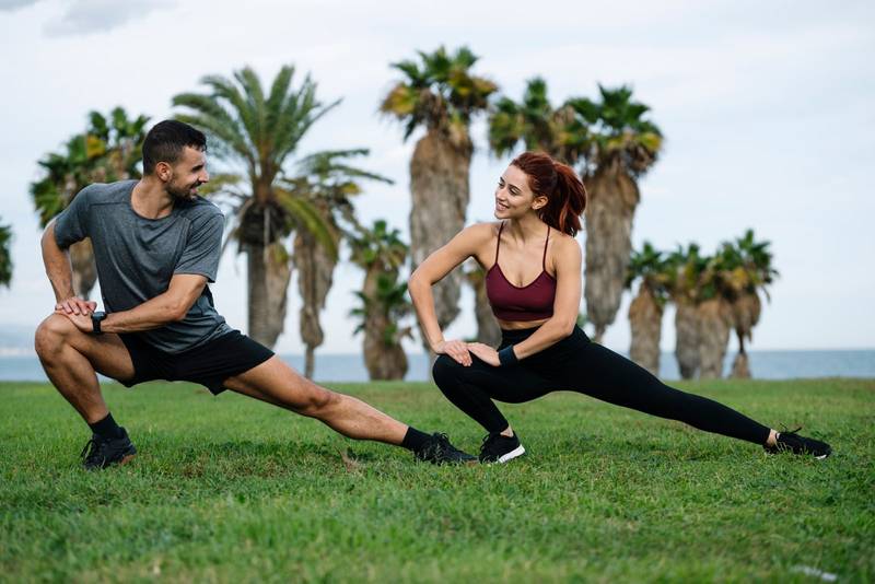 Smiling satisfied athletic male and female sitting in the grass in sportswear training workout outside. Happy young adult couple stretching in the floor after running together in the park. 