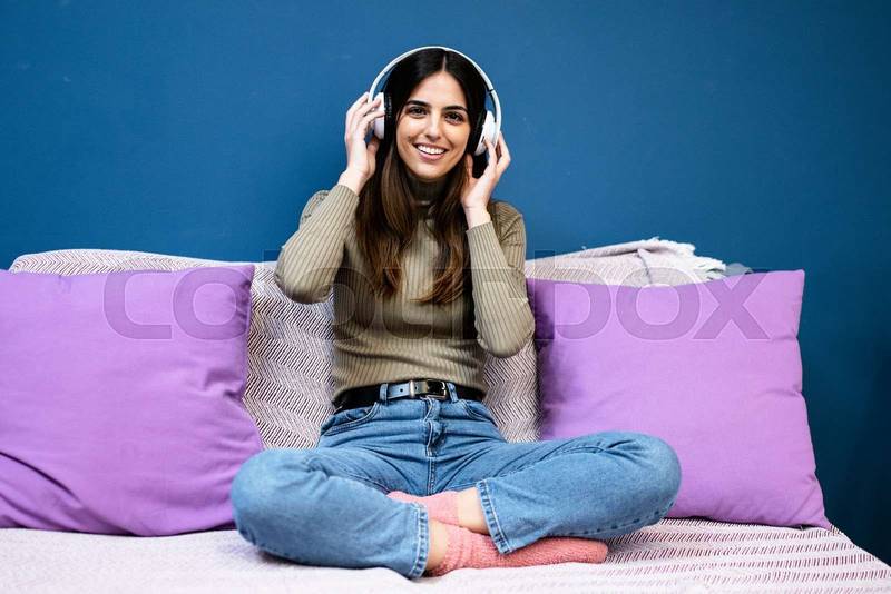 Cheerful young woman listening to music and smiling sitting on couch in livinig room. Happy young girl having fun at home.