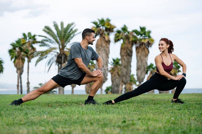 Joyful satisfied and athletic male and female sitting in the grass in sportswear training workout outside. Lovely young adult couple stretching in the floor after running together in park. 