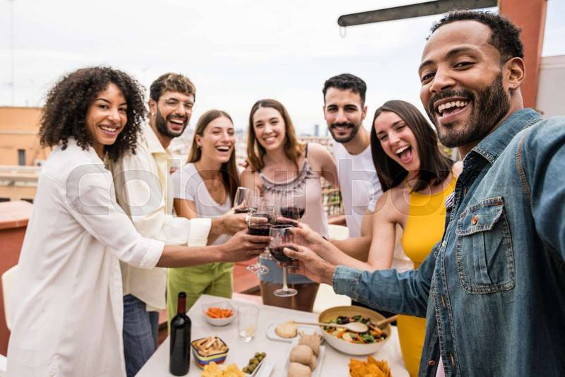 Happy group of friends toasting with wine glasses in a terrace looking at camera. Diverse group of cheerful young people drinking and celebrating at a rooftop dinner Focus on man..