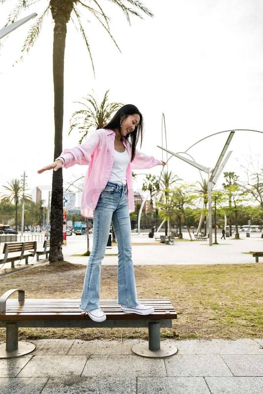 Young happy woman balancing on bench in the street. Carefree and relaxed lady standing on bench park with her arms outstretched in a city.