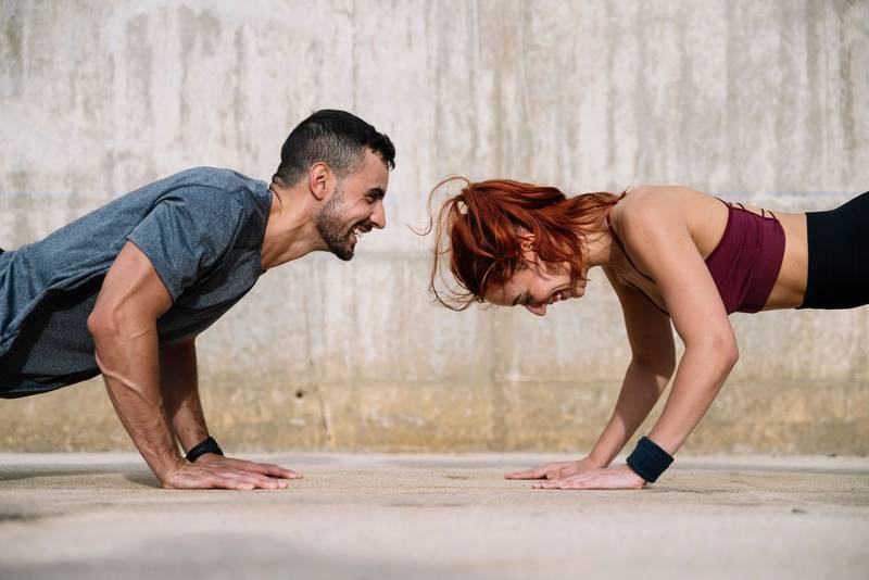 Young adult couple exercising in the floor face to face outside. Joyful athletic male and female doing push ups in the street in sportswear training workout outside. 