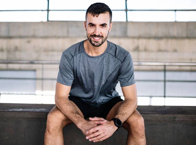 Young athlete man sitting smiling and looking at camera. Happy active runner resting satisfied from training fitness at gym.