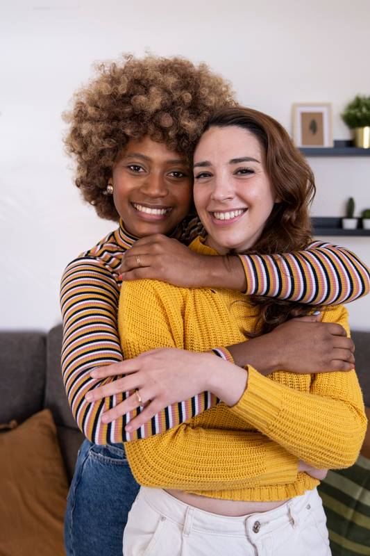 Close up of beautiful and cheerful multiracial lesbian couple hugging and looking at the camera smiling. lovely and happy homosexual women embracing at home staring at the camera.