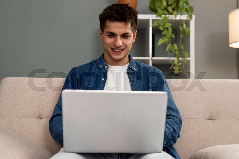Handsome relaxed male student working with laptop while sitting on the couch. Smiling young carefree man using computer at home.