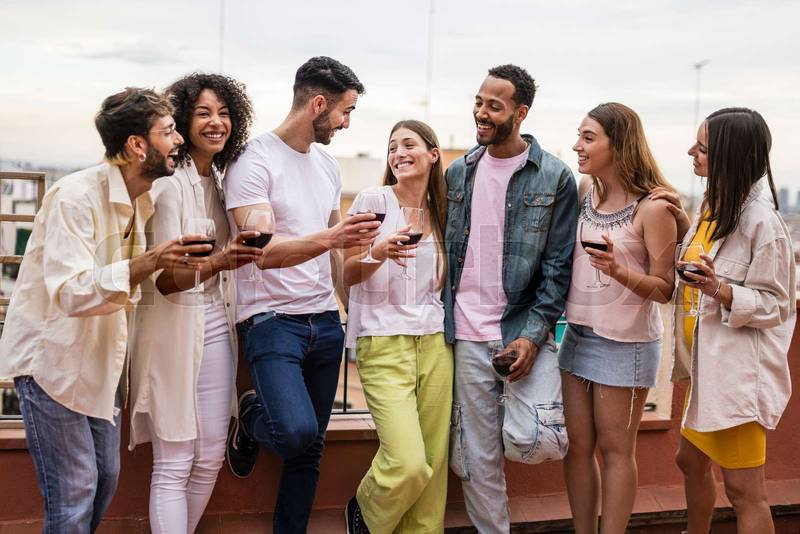 Multiracial group of friends having drinks in a outdoors party standing in a rooftop. Diverse young happy group of millennials smiling and drinking wine in a terrace.