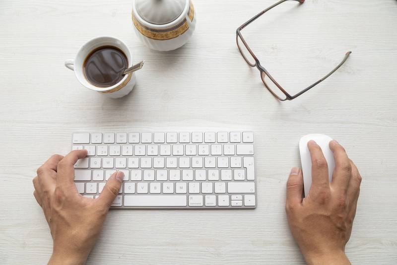 hands of a person typing on a modern keyboard and using a mouse, around them a pair of glasses, a cup of coffee