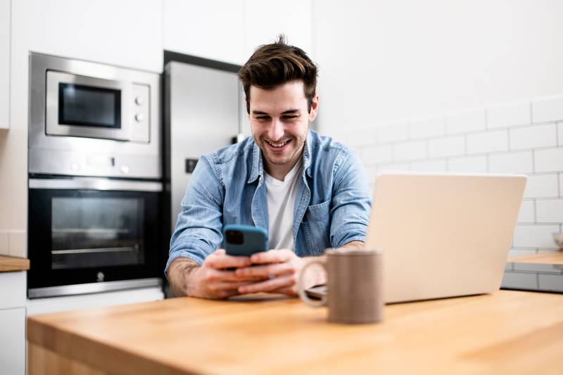 Handsome young man working with a laptop and smiling at the kitchen with a cup of tea - Cheerful, success and business concept