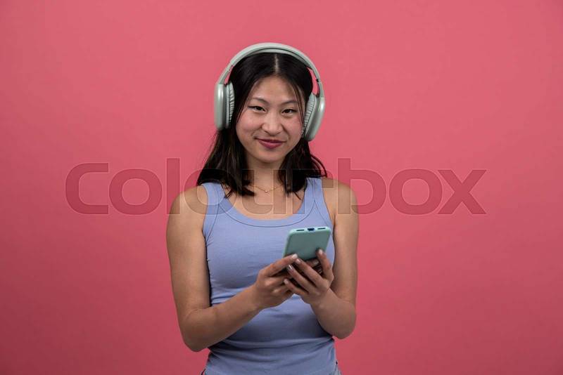 Joyful and satisfied woman with headphone texting with phone looking to the camera. Smiling young female listening to music and using online smartphone isolated on a red background.